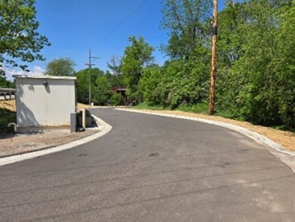 Photo of newly-paved road with a small building on the left side and shrubs and trees on the right.