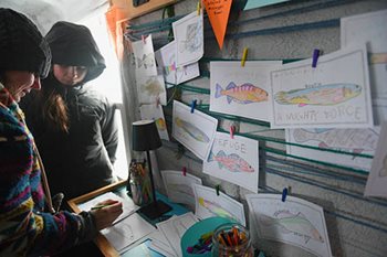 A woman colors a fish near several lines of string with fish pictures attached to them.