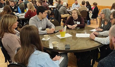 Groups of six to eight people sit at tables in a large meeting room.