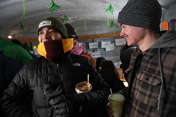 Two people bundled up in their winter gear smile as they explore the shanty with cups in hand.